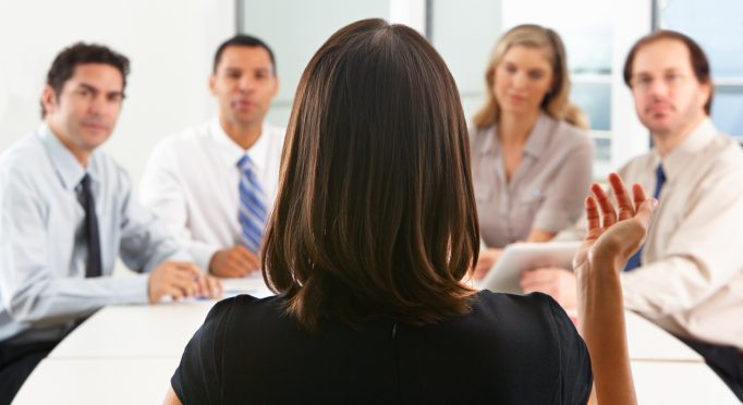 View From Behind As CEO Addresses Meeting In Boardroom Raising Arm Having A Discussion