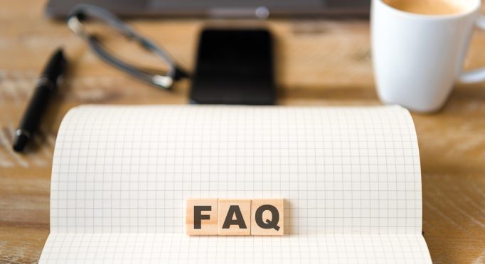 Closeup on notebook over wood table background, focus on wooden blocks with letters making FAQ text. Concept image. Laptop, glasses, pen and mobile phone in defocused background